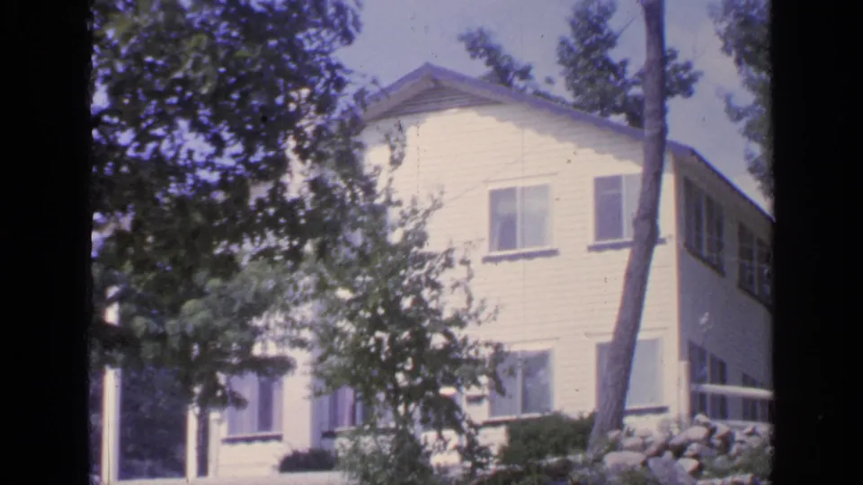1960s home-movie view of a white Greenwood Lake cottage, partly hidden by branches, with stonework and porch railing below.