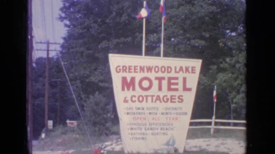 Greenwood Lake Motel & Cottages sign beside a wooded road, with flags, utility poles, and trees visible in the 1964 footage.