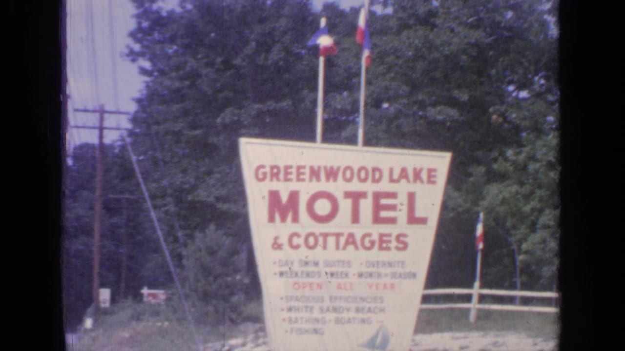 Greenwood Lake Motel & Cottages sign with two flags above it beside trees and a utility pole.