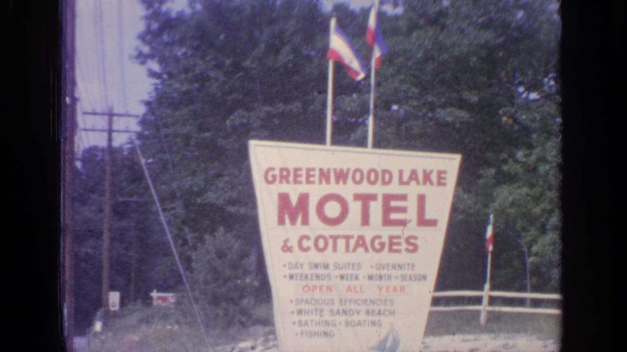 The Greenwood Lake Motel & Cottages sign fills the frame, with small flags above and a wooded roadside background.