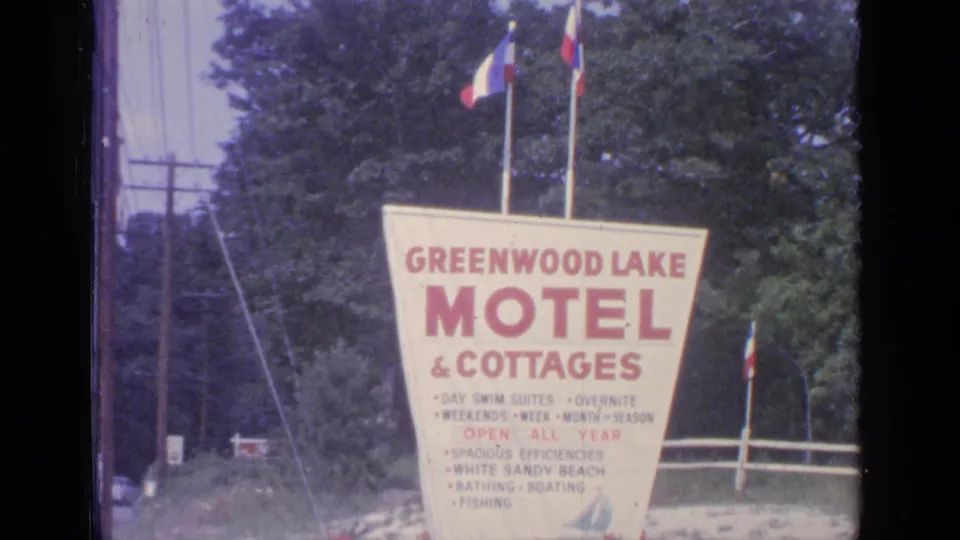 1964 home-movie view of the Greenwood Lake motel sign, showing red lettering, flags, trees, and roadside utilities.