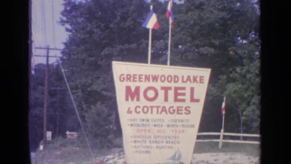 Final 8mm frame holds on the Greenwood Lake Motel & Cottages sign, with flags overhead and a wooded New York roadside behind.