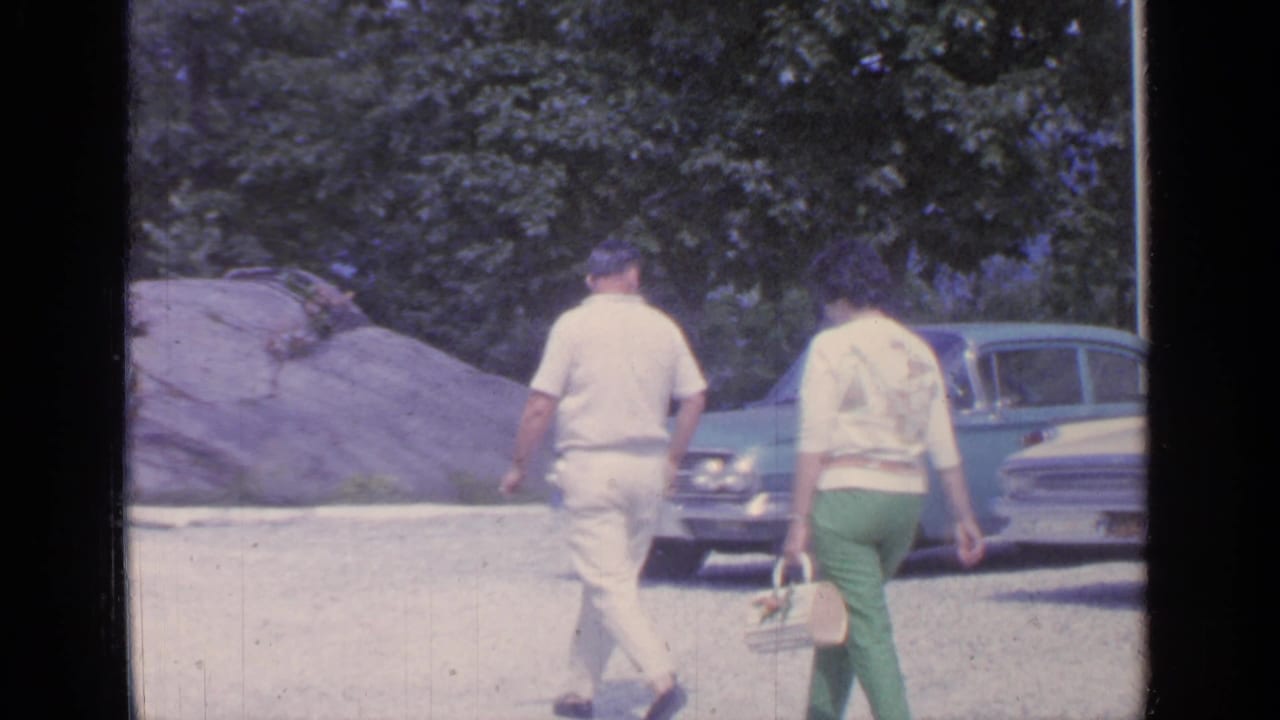 Two adults walk past rocks and a parked blue sedan near trees.