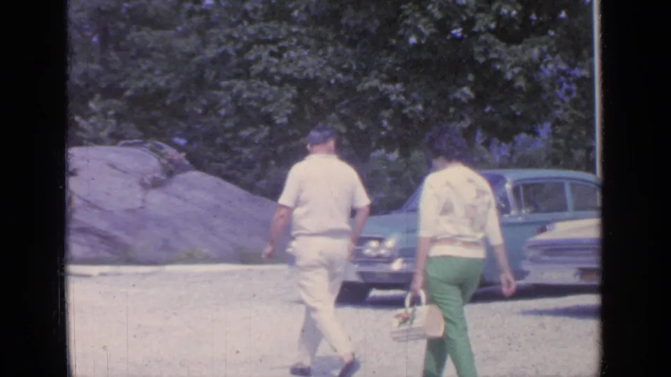 In Greenwood Lake, New York, two adults walk away from the camera near a classic sedan, rocks, and trees in 1964 home-movie footage.