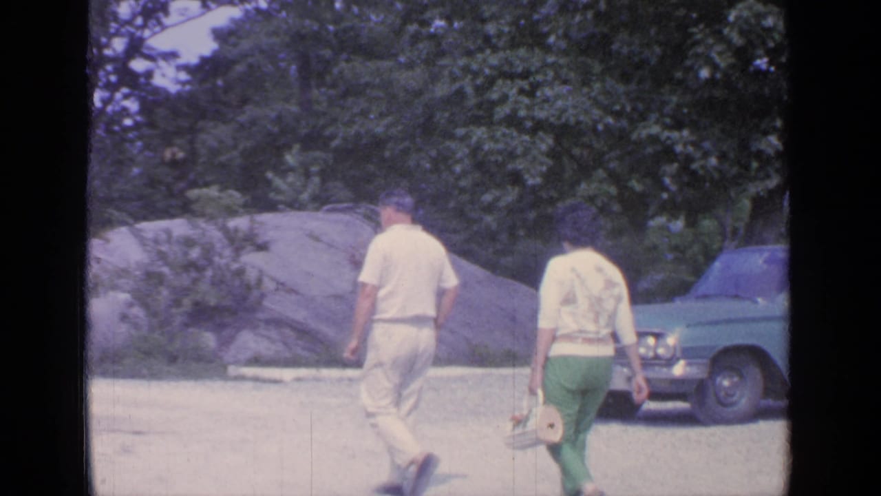 The couple walks toward the motel area with a blue 1960s sedan at right and large rocks and trees behind them.