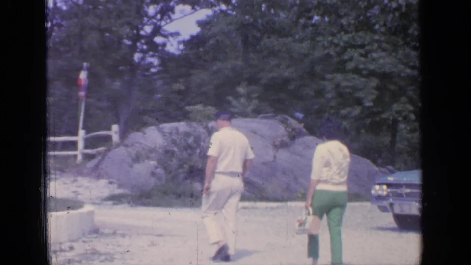 The man and woman move farther from the camera toward a low white fence, with large rocks, trees, and part of a parked car visible.