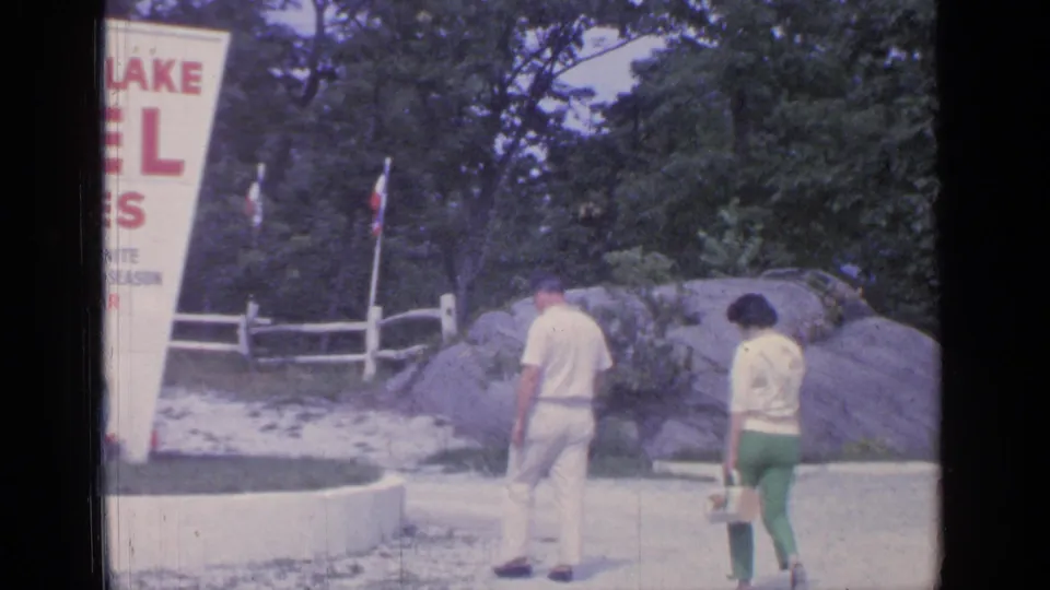 The couple walks near the Greenwood Lake motel sign, with rocks, trees, a white fence, and gravel visible in the 1964 8mm frame.