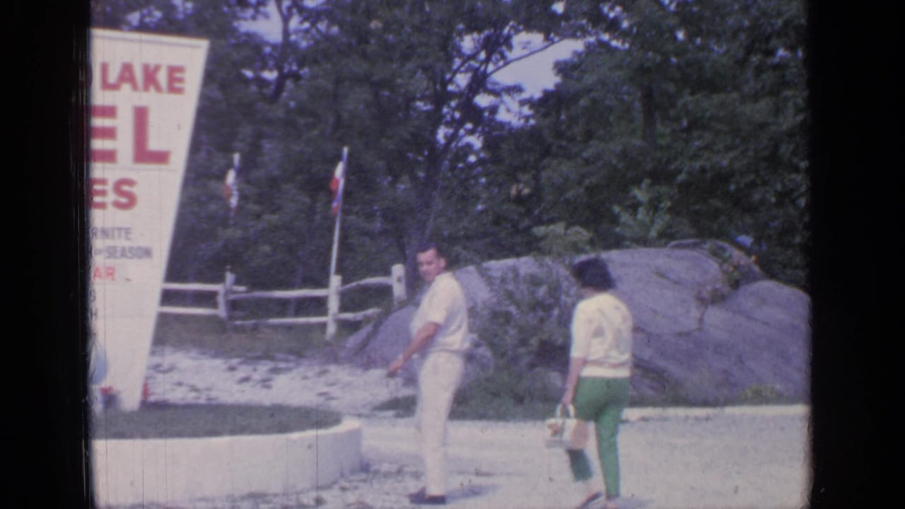 A couple walks near a large white motel sign beside trees, rocks, and a low curb.
