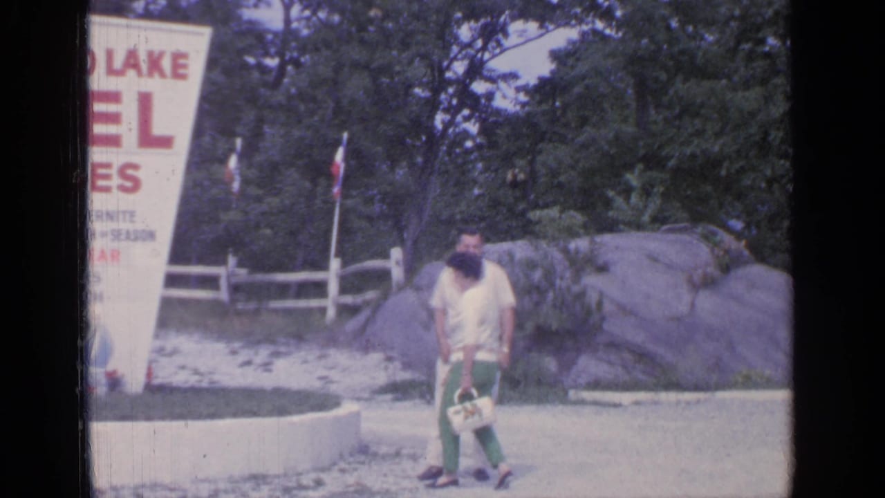 The couple walks closer to the camera with the motel sign partly visible at left.