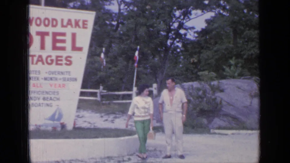 The couple passes the Greenwood Lake Motel Cottages sign, with red lettering, a low curb, flags, trees, and a rock feature visible.