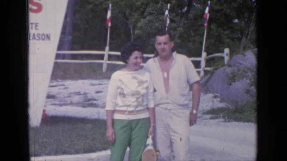 Close 1964 Greenwood Lake frame of the couple near the motel sign, with the woman carrying a small bag and trees, fence, rocks, and flags behind them.