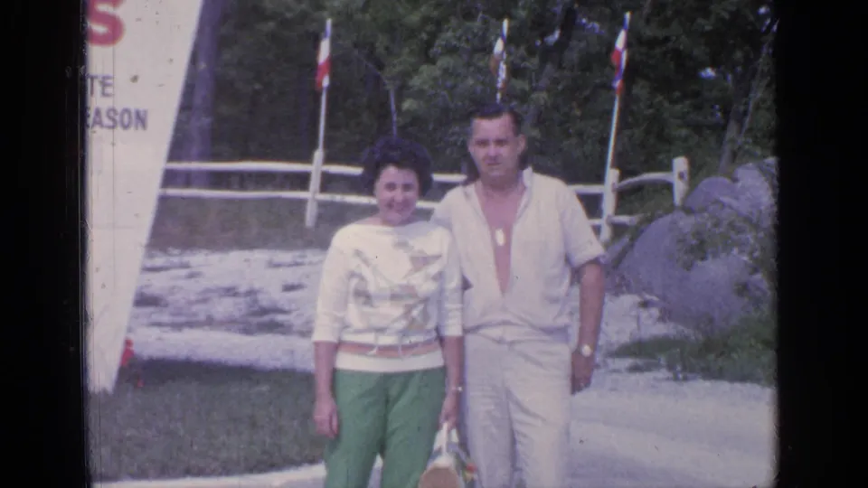 In 1964 Greenwood Lake footage, an adult couple walks toward the camera in casual 1960s clothing near a partial white sign and fence.
