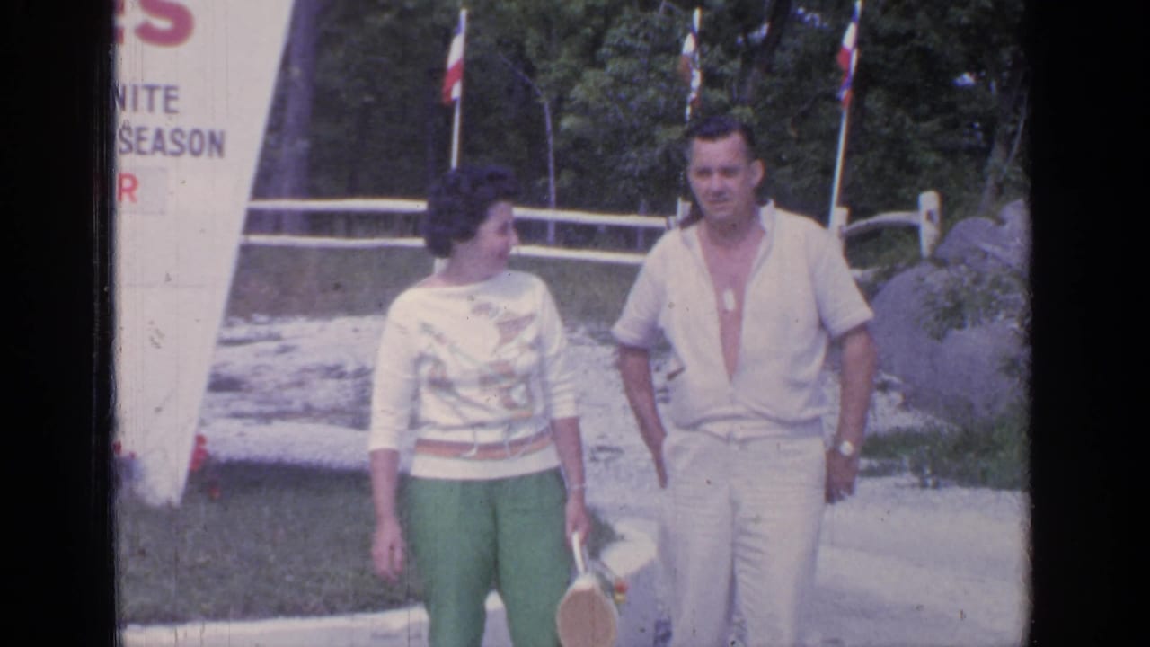 The couple continues walking in the Greenwood Lake home movie, with the woman turning toward the man near a sign, fence, and trees.