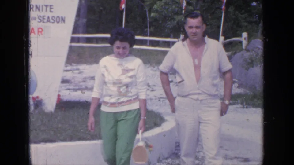 A closer 1964 Greenwood Lake frame shows the couple walking toward the camera, with bright green pants, white clothing, lawn, curb, and partial signage visible.