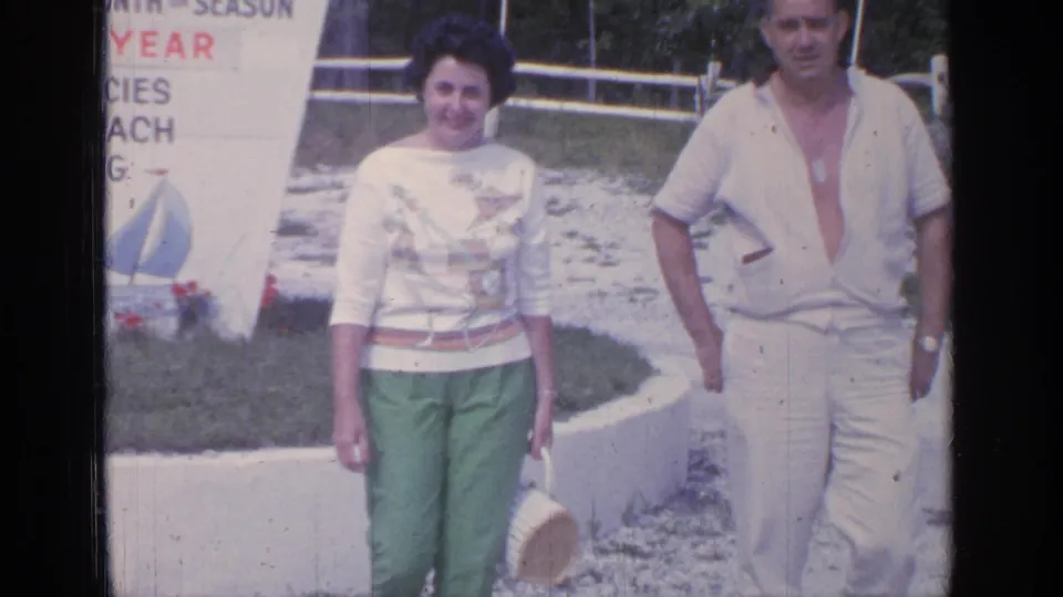 The final frame shows the smiling woman and man in 1960s attire walking past partial seasonal text, with fence rails, lawn, and trees behind them.