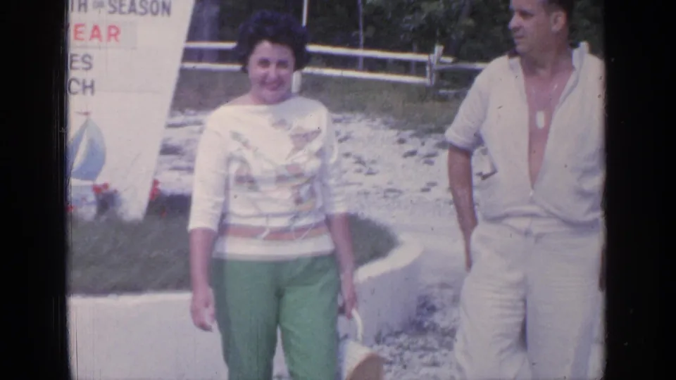 A couple in 1964 Greenwood Lake footage walks near a seasonal beach sign, with the woman carrying a basket and the man in white clothing.