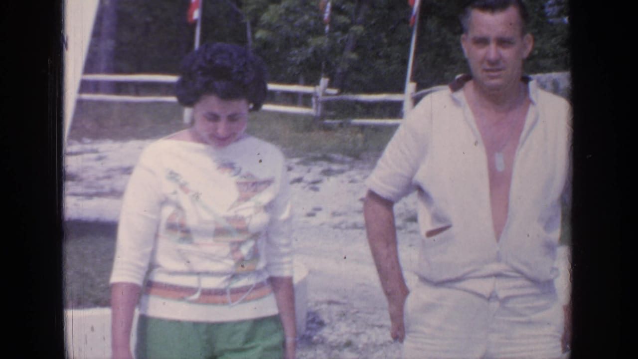 The Greenwood Lake couple walks closer to the camera, with a beach-area sign, white fence, and trees visible behind them.