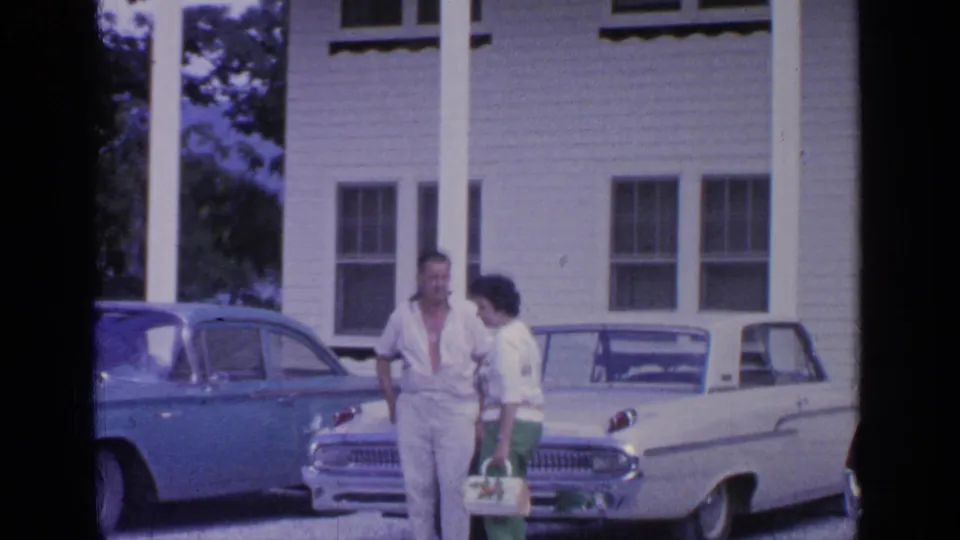 The couple pauses near two classic cars outside a large white columned building in the Greenwood Lake resort footage.