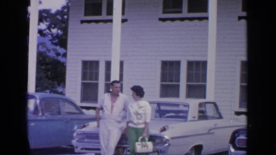 The couple walks together in front of a white columned building, with blue and white classic cars parked nearby.
