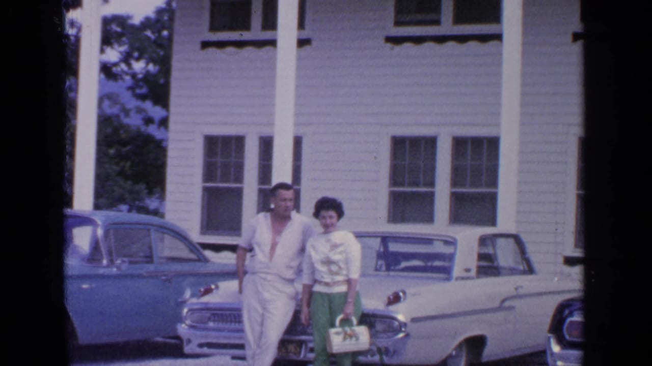 Couple standing beside classic cars in front of a white clapboard house.