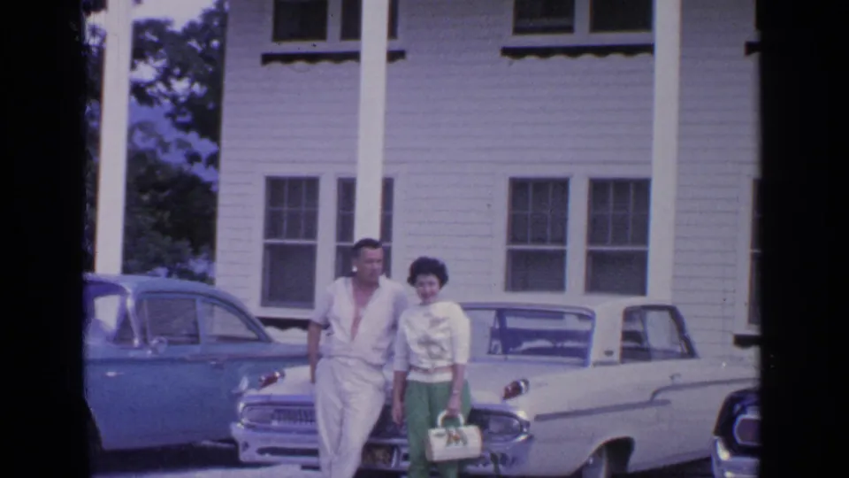 In Greenwood Lake, New York, a couple poses near a white convertible and blue sedan outside a white clapboard house in 1964 footage.