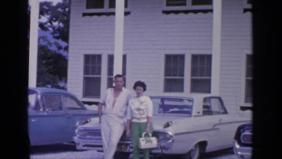 The couple remains posed in front of the house, with the white convertible and blue sedan framing the 1964 Greenwood Lake scene.