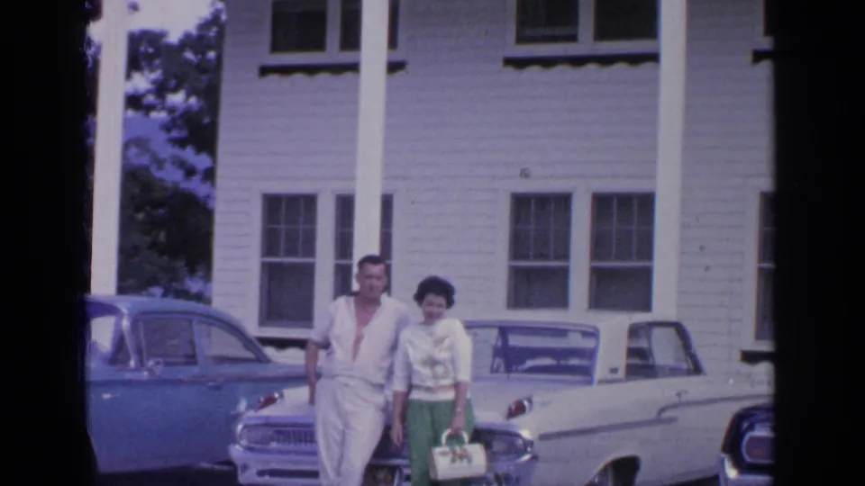 The couple stands close together outside the white house, with the blue sedan at left and the white convertible behind them.