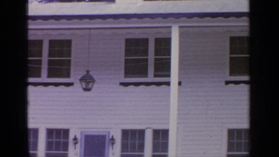 1964 home-movie frame of a white two-story Greenwood Lake house exterior with columns, windows, door, and porch lights.