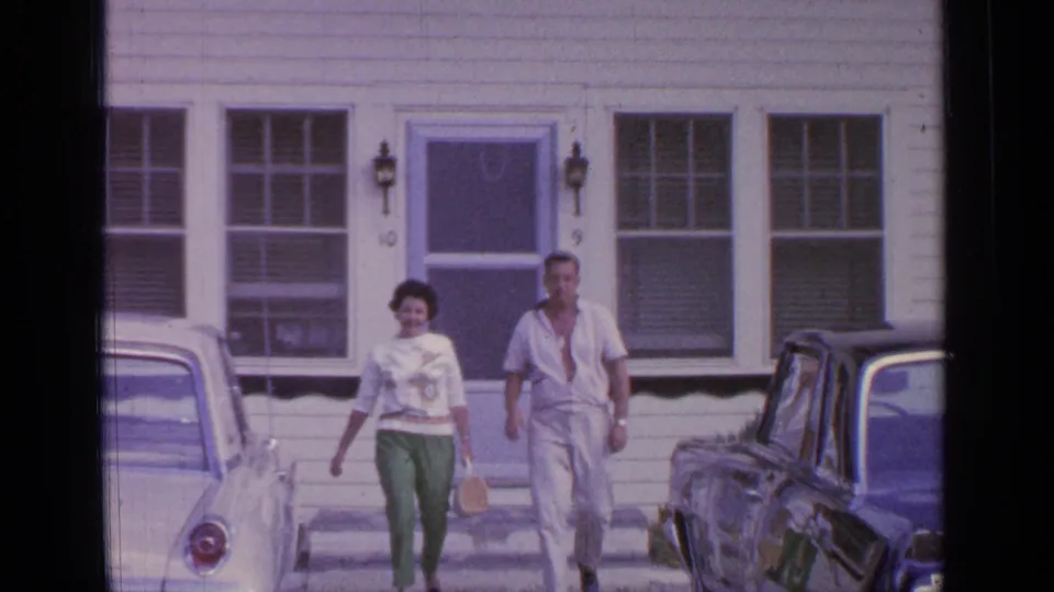 1964 Greenwood Lake frame showing a woman and man walking toward the camera from a white house, with parked cars nearby.