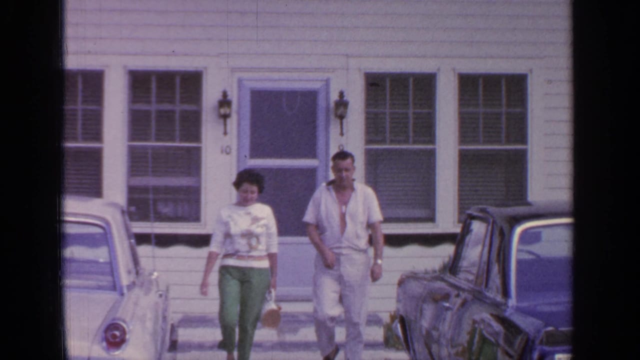 Two adults walk from a white clapboard house between parked cars.