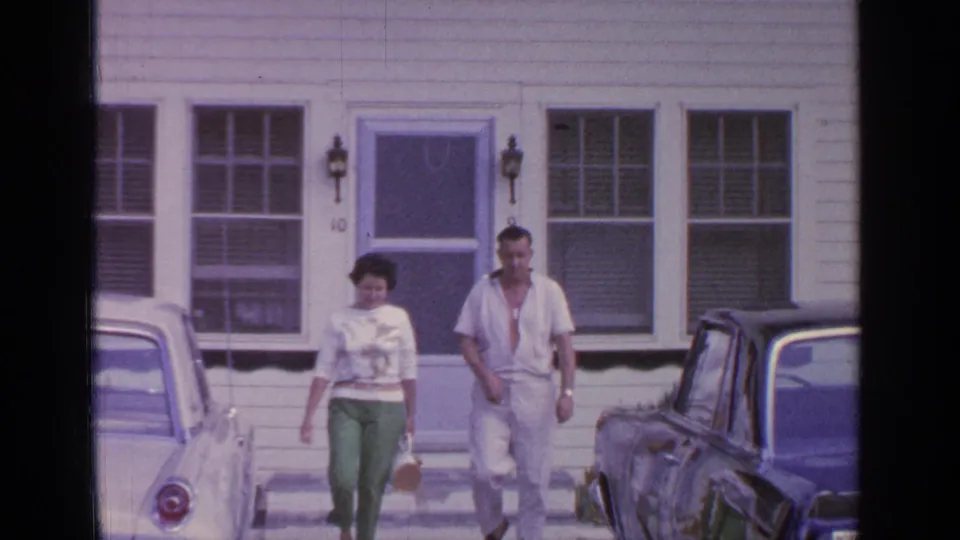 In 1964 Greenwood Lake home-movie footage, a couple walks from a white clapboard house toward parked mid-century cars.