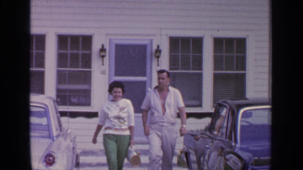 A woman and man walk forward from the house, with cars on both sides.