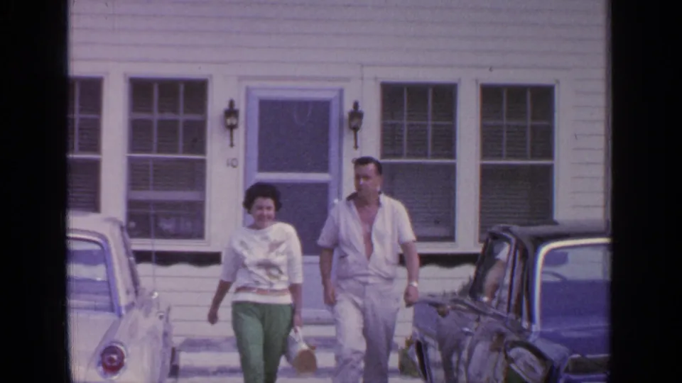 The couple continues down from the Greenwood Lake house, the woman in green pants and the man in light clothing near parked cars.