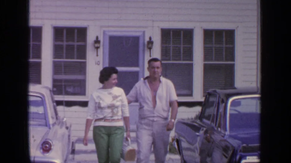 The woman glances toward the man as they walk together outside the 1964 Greenwood Lake house, with cars flanking the path.
