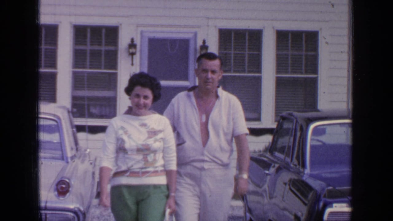 Couple walking toward the camera between parked cars outside a white clapboard building.