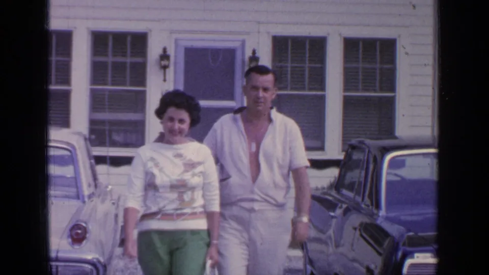 In supported 1964 Greenwood Lake footage, a couple walks toward the camera outside a white clapboard building with parked cars nearby.