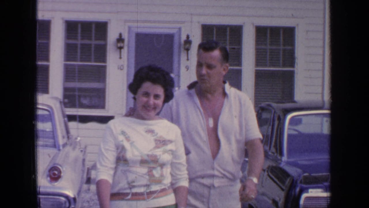 Couple closer to the camera with parked cars and numbered doors behind them.