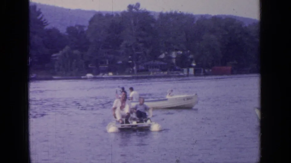 A small group sits on a paddle boat on Greenwood Lake, with trees, shoreline structures, and hills in the distance.