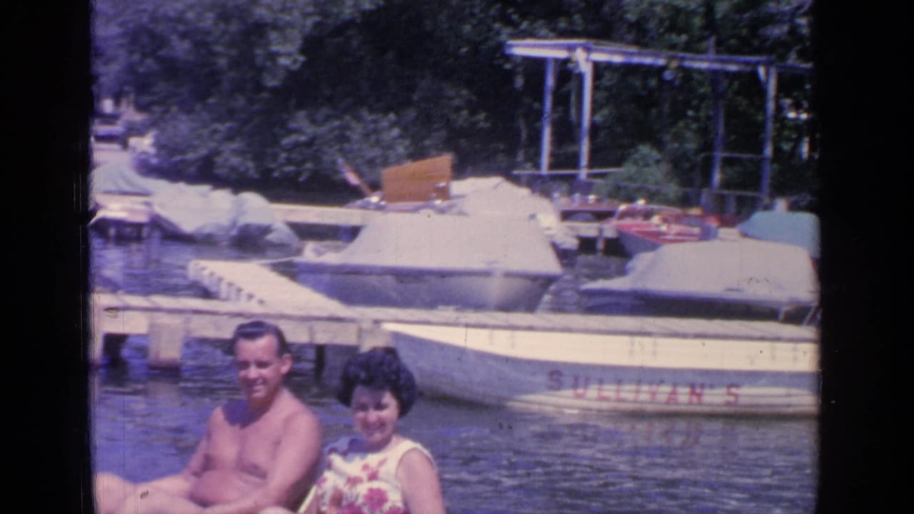 Couple seated on a small boat or float near docks and covered boats on a lake.