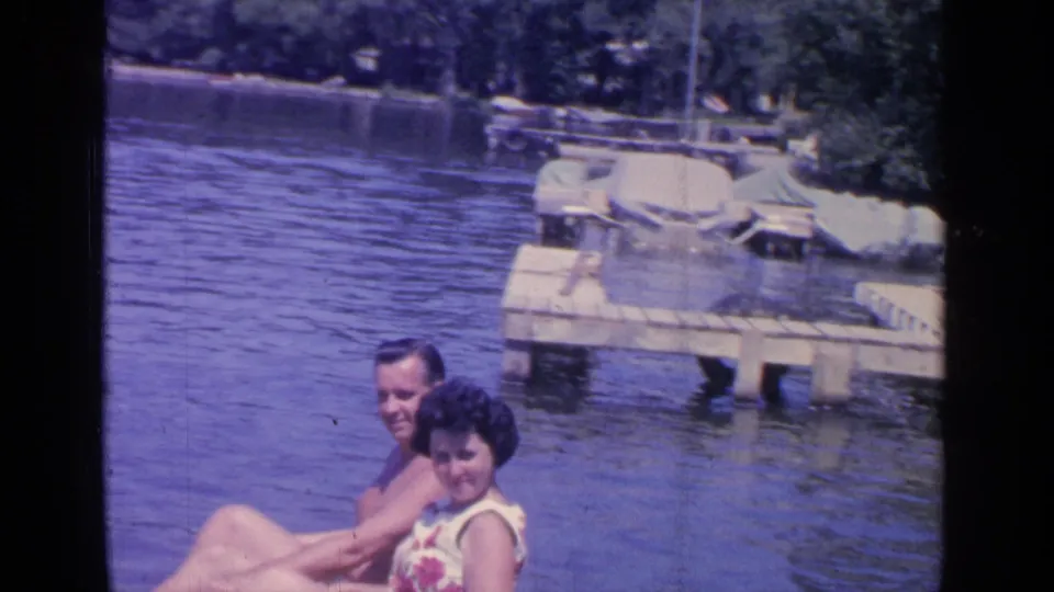 The home-movie frame shows the couple smiling toward the camera on Greenwood Lake, with rippling water, dock planks, and boats behind them.
