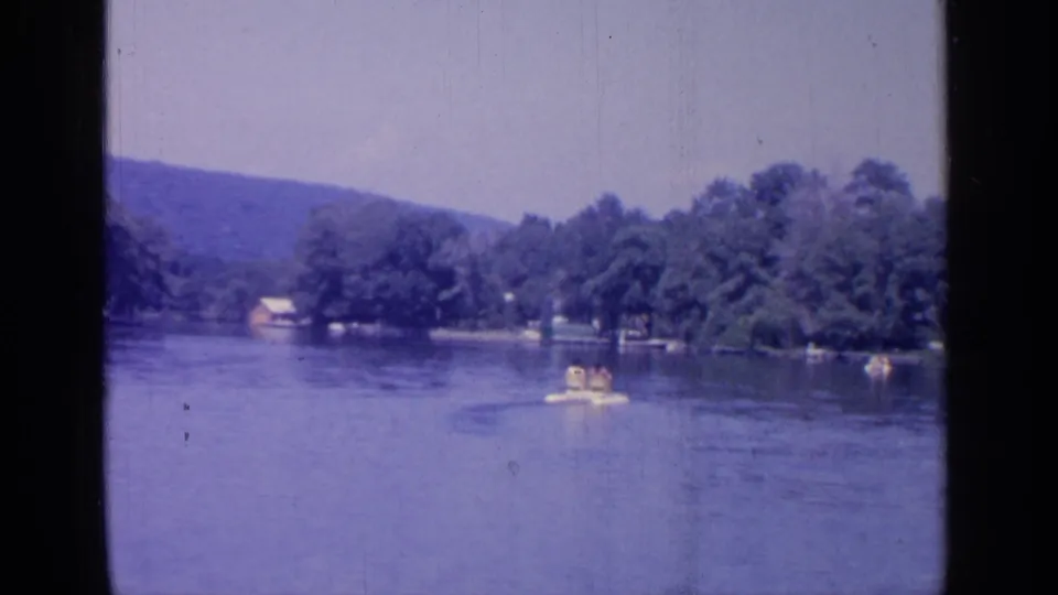 1964 Greenwood Lake home-movie frame showing a small boat with riders on calm water, with trees and low hills beyond.