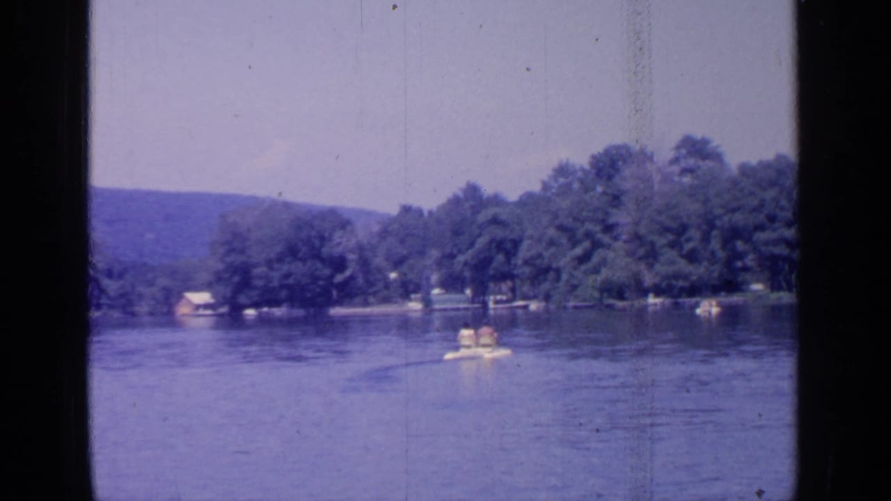 A distant small recreational boat travels on Greenwood Lake in 1964, framed by trees, shoreline, and low hills.