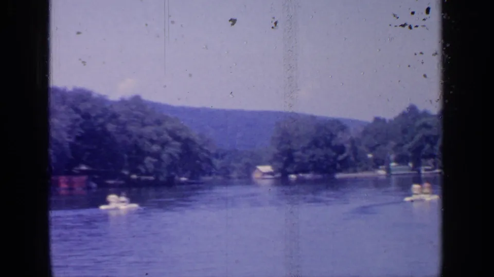 Greenwood Lake 1964 frame with small pleasure boats on the water, wooded shore structures, and hazy hills beyond.