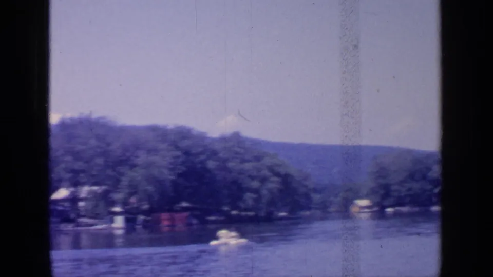 A small white recreational boat crosses Greenwood Lake, with shoreline trees, a few structures, and hills behind it.
