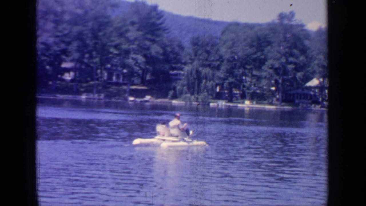 Person paddling a white boat on a lake with trees and hills behind.
