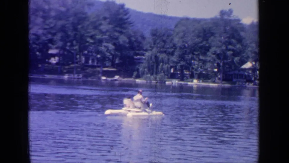 1964 Greenwood Lake home-movie frame showing a person in a white paddleboat on calm water near a wooded shore.