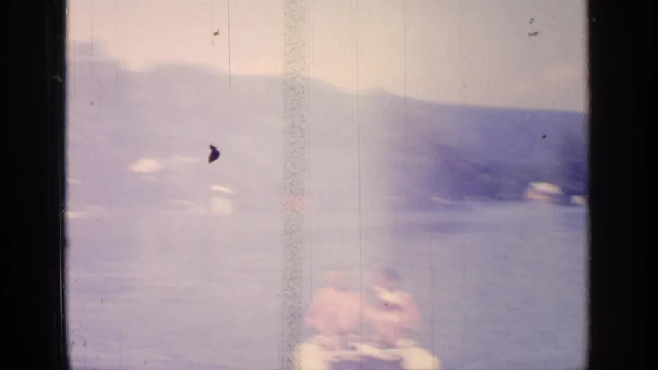 Soft, faded 1964 8mm frame from Greenwood Lake showing two adults in a small paddle boat on open water.