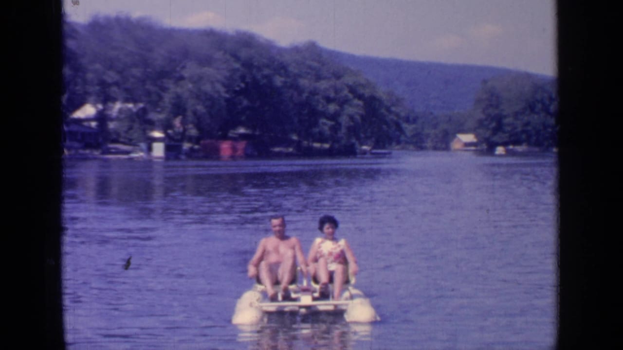 Two adults ride a small paddle boat on a lake near a wooded shoreline.