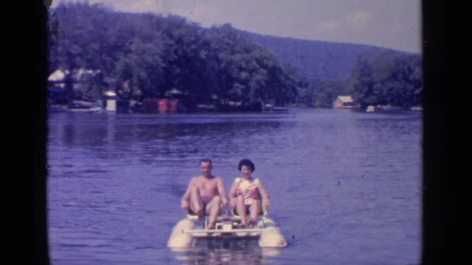 1964 home-movie frame of two adults on a paddle boat at Greenwood Lake, with wooded shore and distant hills visible.