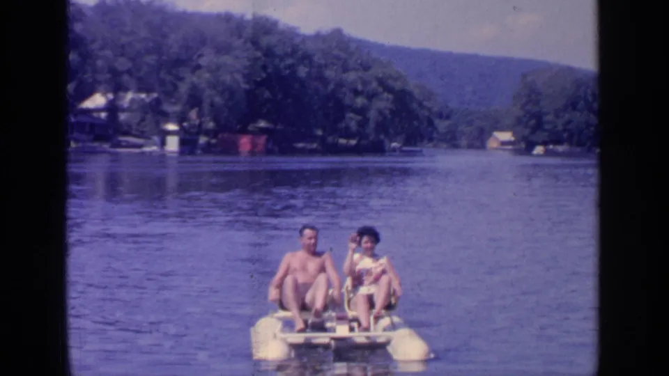Two adults in swimwear sit on a small paddle boat on Greenwood Lake, with wooded hills and lakeside buildings in the background.